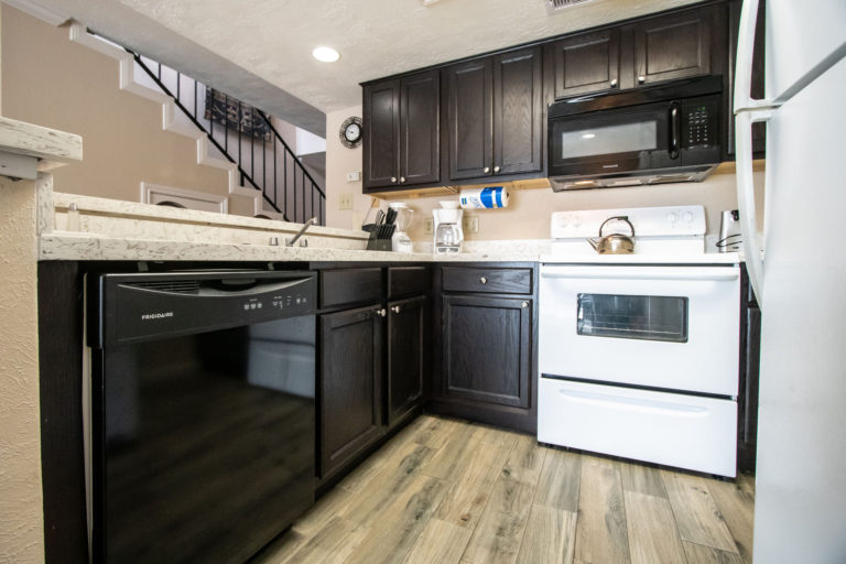 Photo of kitchen with dark wooden cabinets, black microwave, black dishwasher, and white stove. Room 225